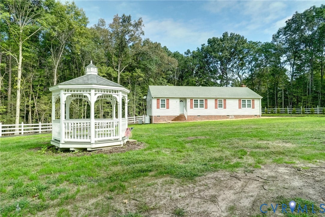 4130 Charles City Road, Charles City, VA 23231 Exterior grounds featuring a white gazebo with a shingled roof, a residence with light-colored siding, a brick foundation, and extensive white rail fencing set against a wooded backdrop