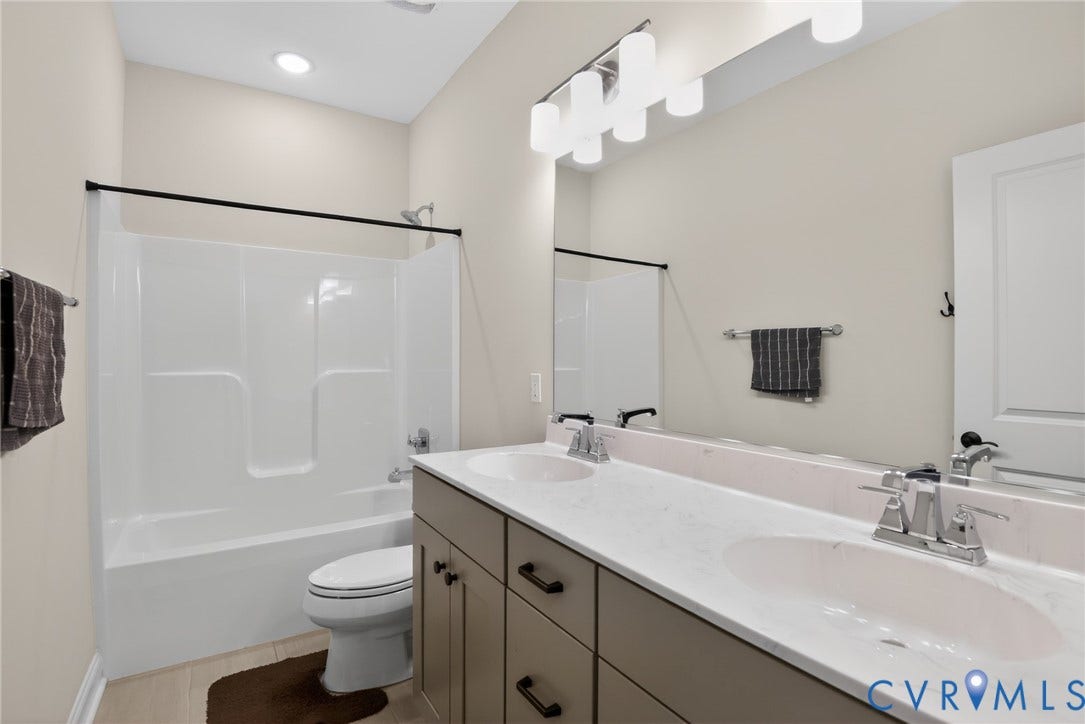 Glen Allen, VA 23059 Bathroom featuring a dual-sink vanity with light-toned stone-surface countertop, white tub-and-shower combination, large wall-mounted mirror, and multi-bulb overhead lighting fixture