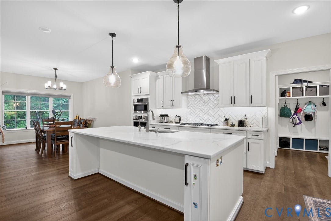 Glen Allen, VA 23059 Kitchen featuring a large central island with quartz countertops, custom white cabinetry, herringbone tile backsplash, stainless steel range hood, and wood-finish flooring