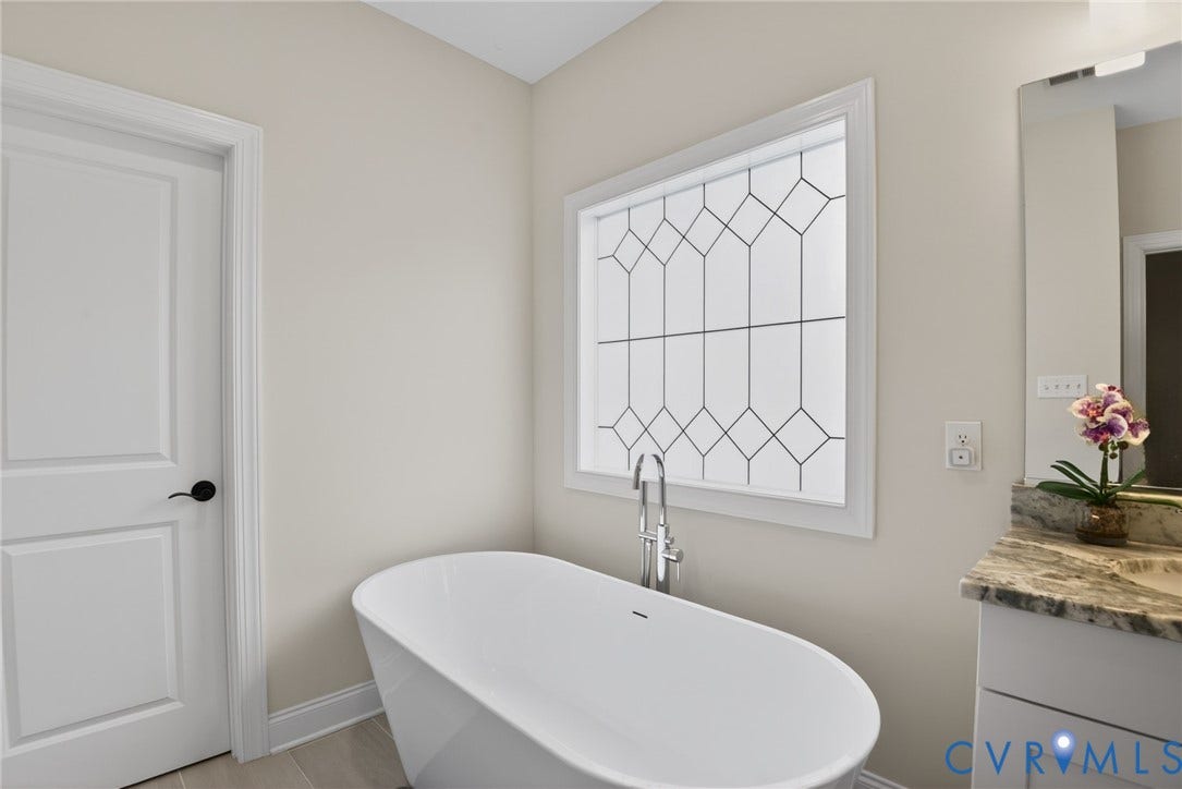 Glen Allen, VA 23059 Freestanding oval soaking tub with floor-mounted chrome faucet, decorative lattice-pattern window, solid surface vanity countertop, and shaker-style cabinetry