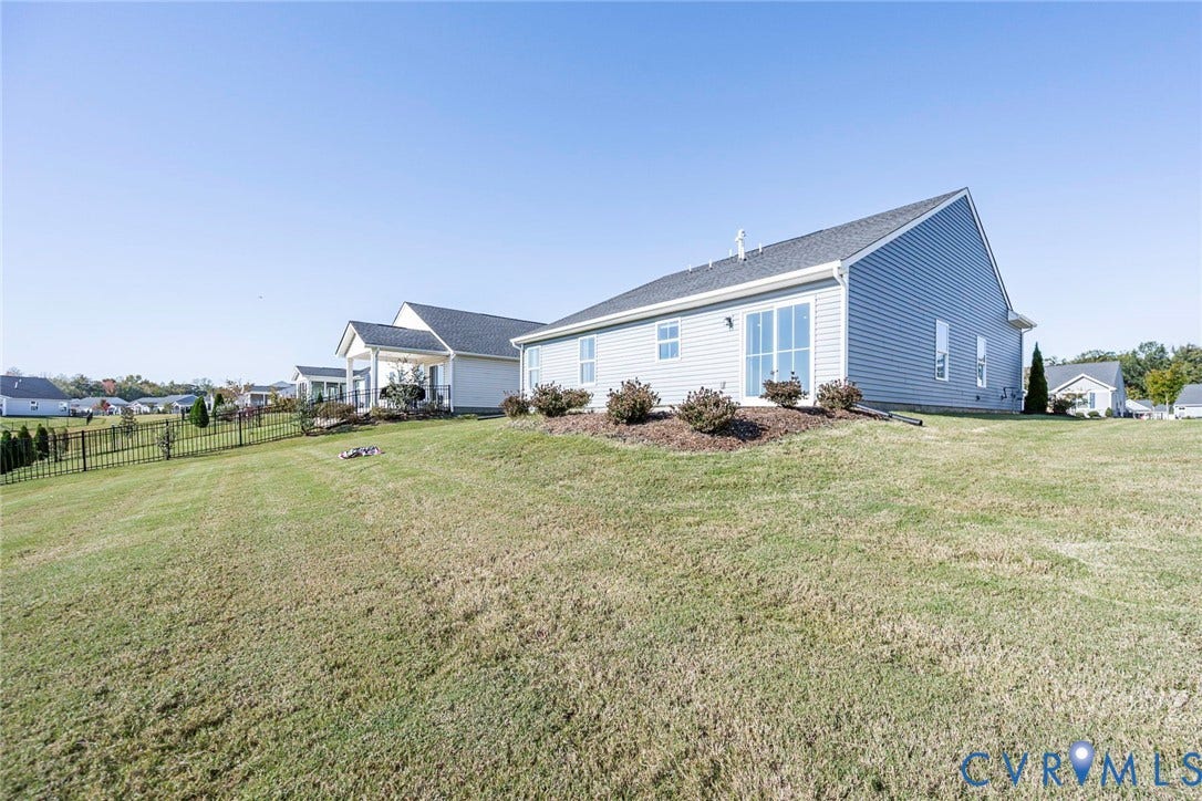 7577 Southamptonshire Way, Autum Pines, New Kent, VA 23124 Exterior of the residence featuring grey horizontal lap siding, a gabled roofline, glass sliding doors, and a manicured lawn enclosed by a metal fence