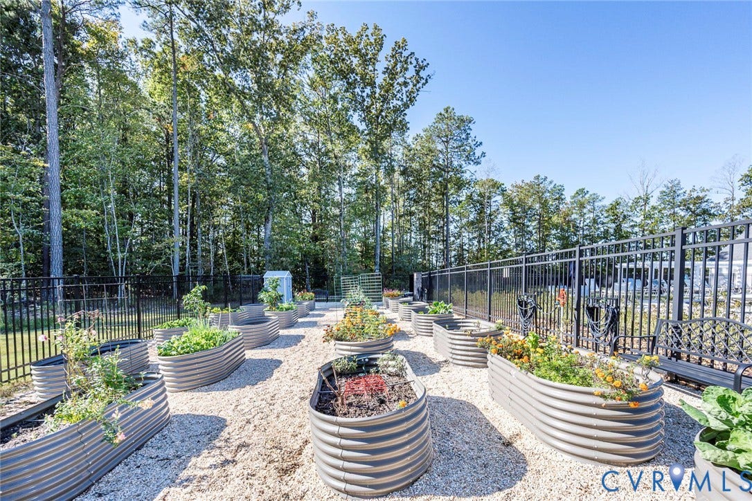 7577 Southamptonshire Way, Autum Pines, New Kent, VA 23124 Garden area featuring multiple corrugated metal raised planting beds set within a gravel-surfaced space, enclosed by black metal fencing and bordered by mature trees