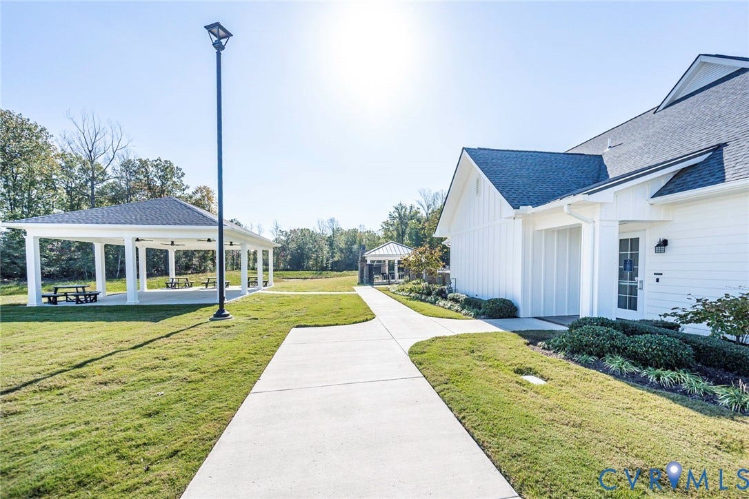 7577 Southamptonshire Way, Autum Pines, New Kent, VA 23124 White board-and-batten siding with a dark asphalt shingle roof, concrete walkways, expansive manicured lawn, and a covered open-air pavilion