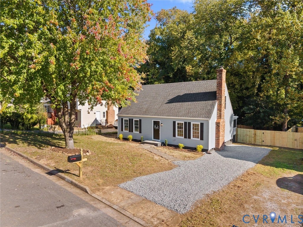 5105 Carryback Drive, Richmond, VA 23234 Cape Cod-style residence featuring grey siding, dark shutters, a brick chimney, a gravel driveway, and a wood-paneled privacy fence
