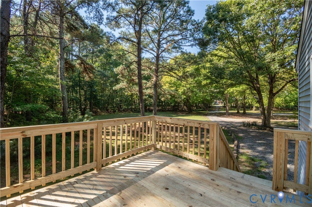 9045 Blounts Corner Road, Smithfield, VA 23430 Elevated wood deck featuring vertical baluster railings and stairs leading to the grounds, accented by mature trees and a gravel driveway
