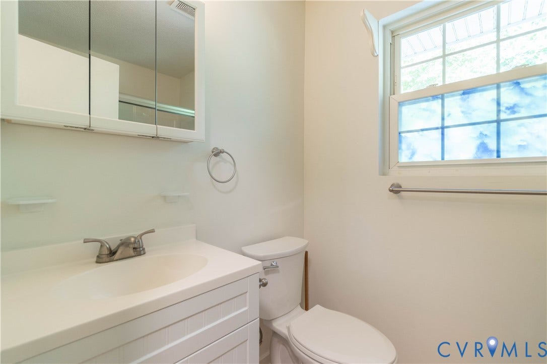 9045 Blounts Corner Road, Smithfield, VA 23430 White vanity with integrated basin and metallic faucet, mirrored medicine cabinet, wall-mounted towel ring, toilet, and single-hung window