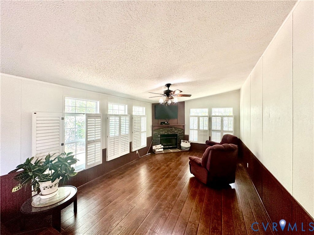 25400 SAYLERS CREEK ROAD, Deatonville, Jetersville, VA 23083 Living space featuring wood-finish flooring, corner fireplace with stone-surround facade, multiple windows with attached plantation shutters, and a ceiling-mounted light fixture with fan