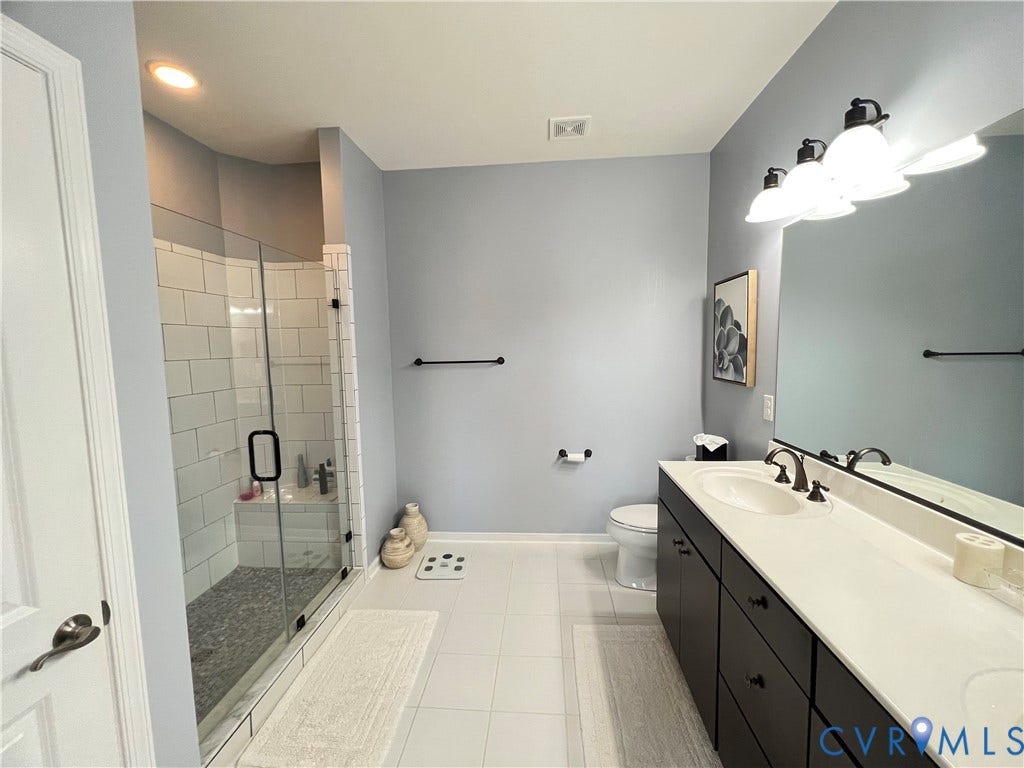 2310 Liesfeld Parkway, West Broad Village, Henrico, VA 23060 Bathroom featuring a dual-sink vanity with a solid-surface countertop, dark cabinetry, a glass-enclosed shower with subway tile surround, and a large wall-mounted mirror with integrated lighting