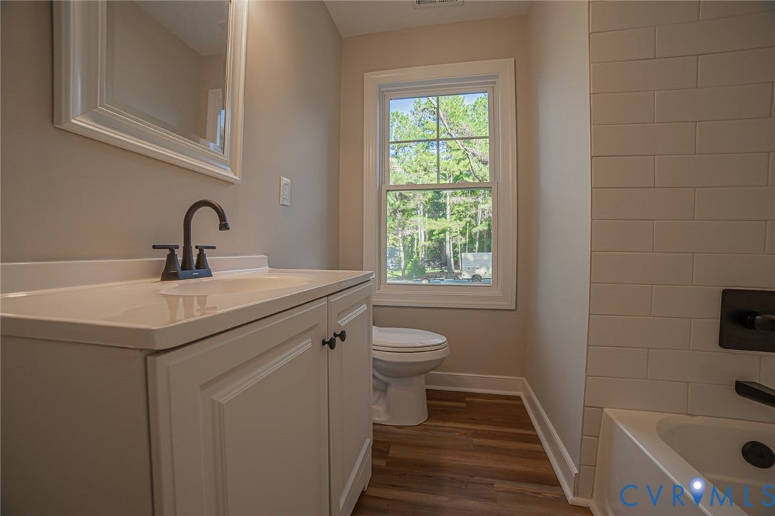 White vanity with a rectangular sink, matte black faucet, subway-tiled tub surround, and wood-finish flooring
