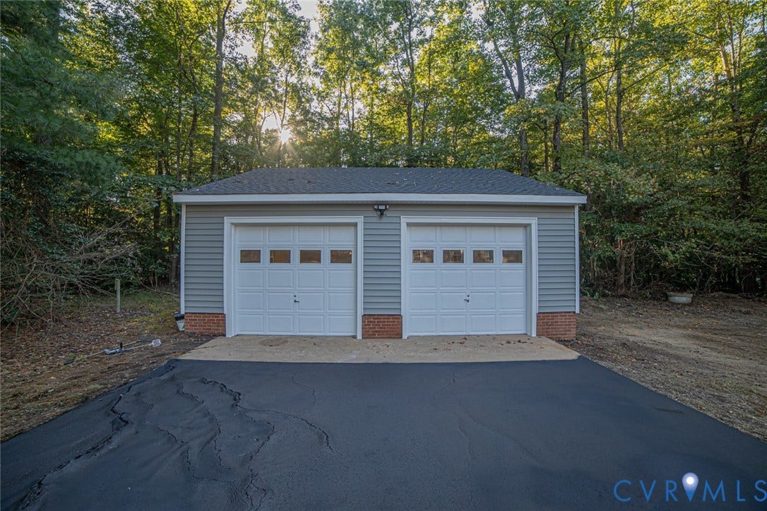Detached two-car garage featuring gray horizontal siding, red brick foundation accents, dual paneled garage doors with upper glass inserts, and an asphalt driveway