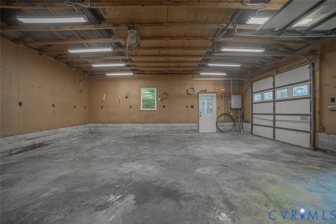Spacious garage featuring a concrete floor, exposed wood beam ceiling, multiple overhead linear light fixtures, panelled wall surfaces, and a multi-panel garage door