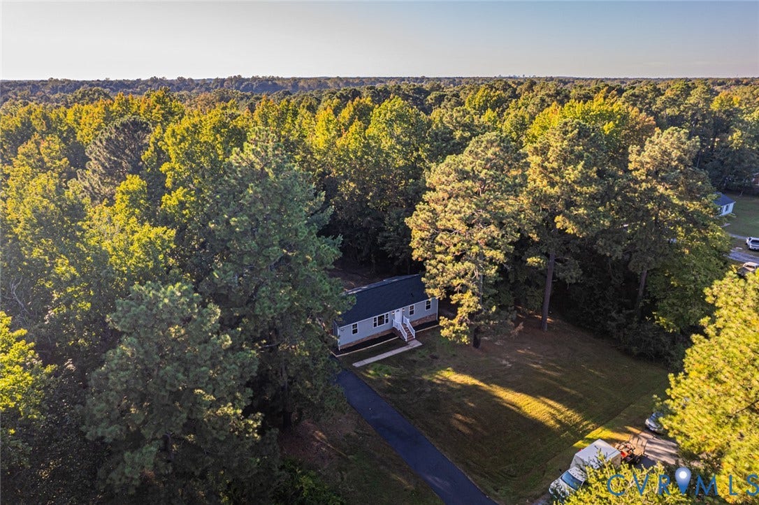 Aerial perspective of a detached home featuring a paved driveway, open lawn, and dense mature tree coverage