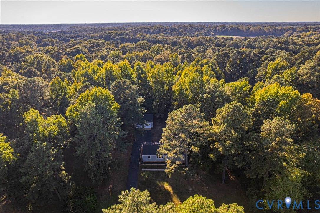 Aerial perspective of a detached residence featuring a paved driveway, multiple structural components, and expansive surrounding woodland