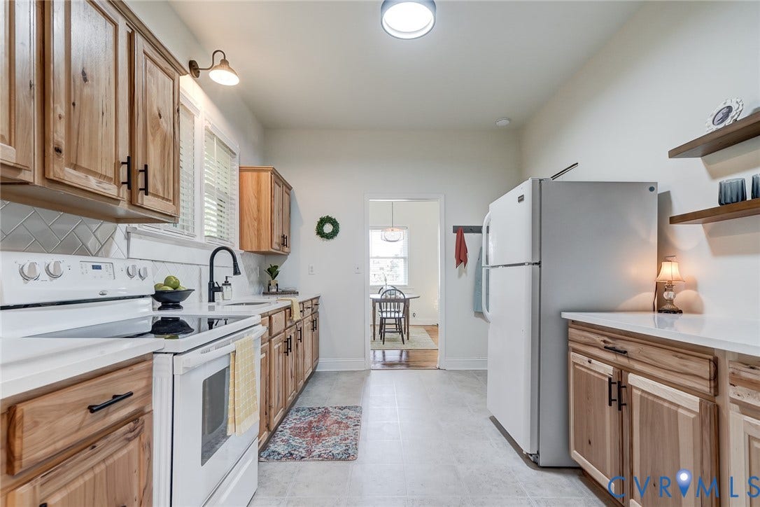 Kitchen featuring natural wood cabinetry with matte black hardware, light-toned countertops, tile backsplash, and mounted floating shelving