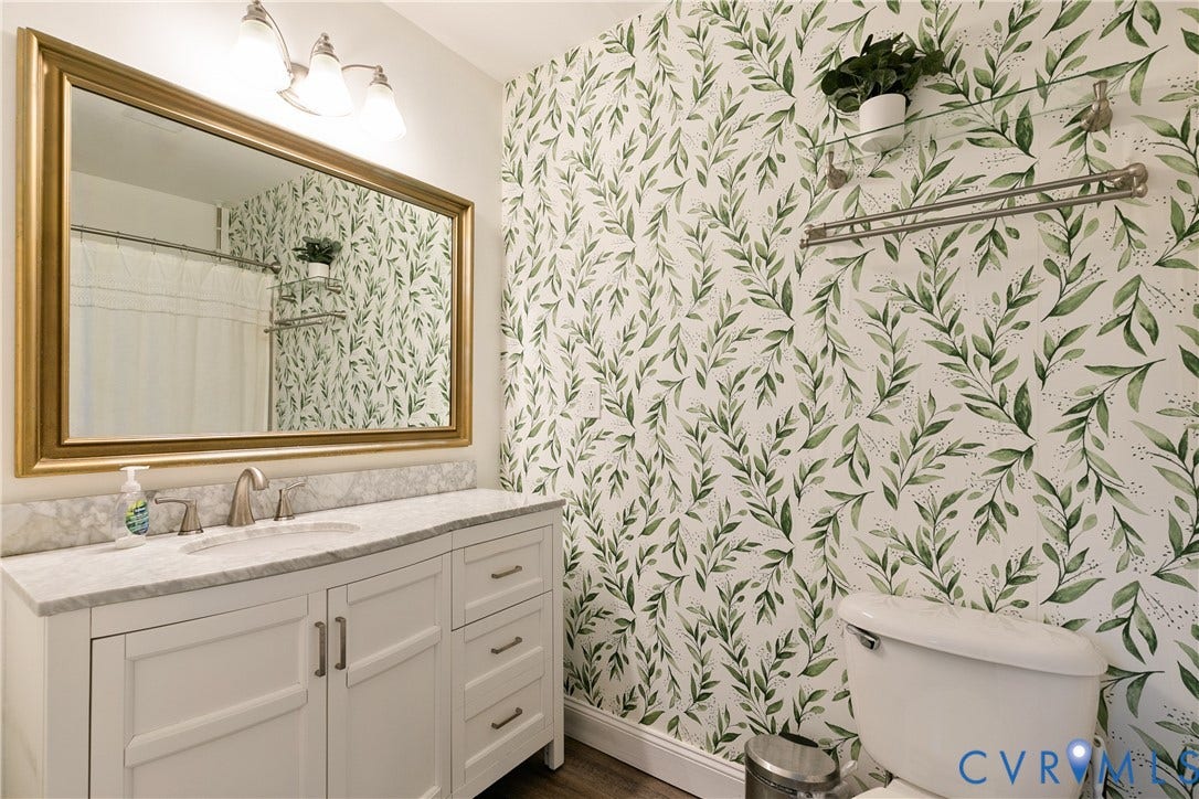 2108 Parkside Avenue, Henrico, VA 23228 White vanity with stone-finish countertop, integrated sink, gold-tone framed mirror, three-light fixture, and wall-mounted metal shelving
