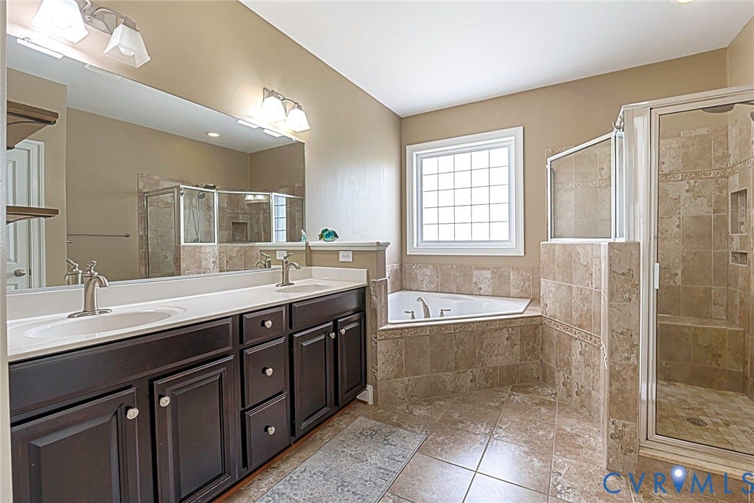 9118 Colonnade Circle, Ashland, VA 23005 Dual-sink vanity with dark-finish cabinetry, solid-surface countertops, corner soaking tub, and enclosed glass-door shower with stone-tile surround