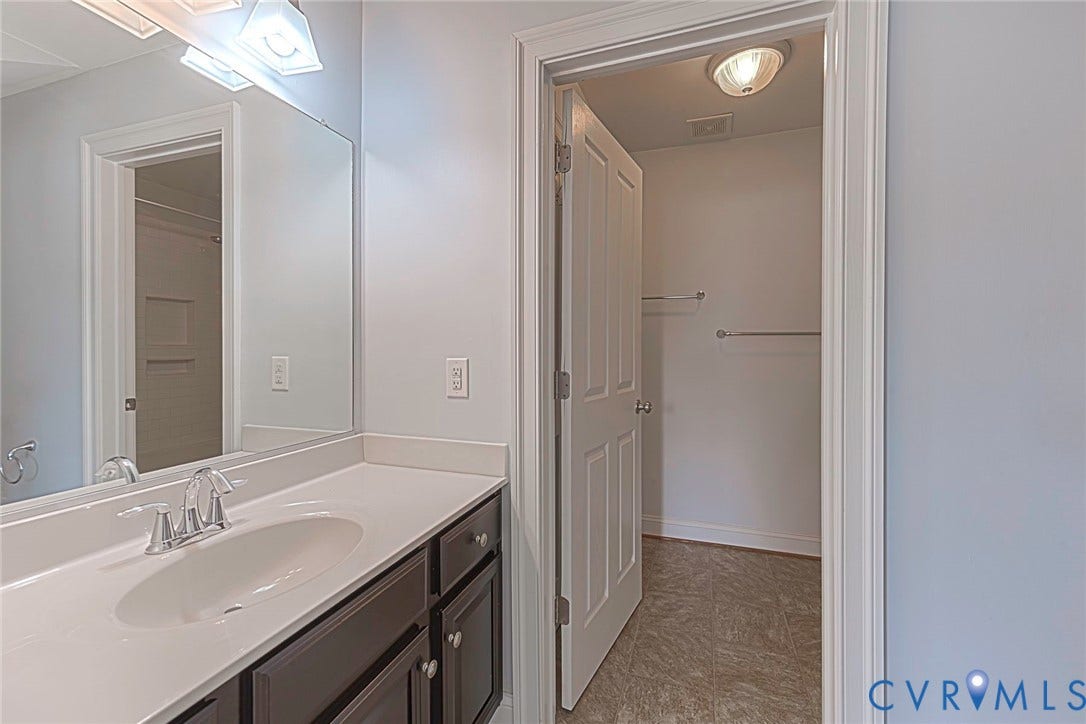 9118 Colonnade Circle, Ashland, VA 23005 Vanity featuring a solid-surface countertop with an integrated oval sink and dark-toned cabinetry, complemented by a large wall-mounted mirror, brushed-finish fixtures, and a six-panel door opening to a storage area with tile flooring and installed hardware