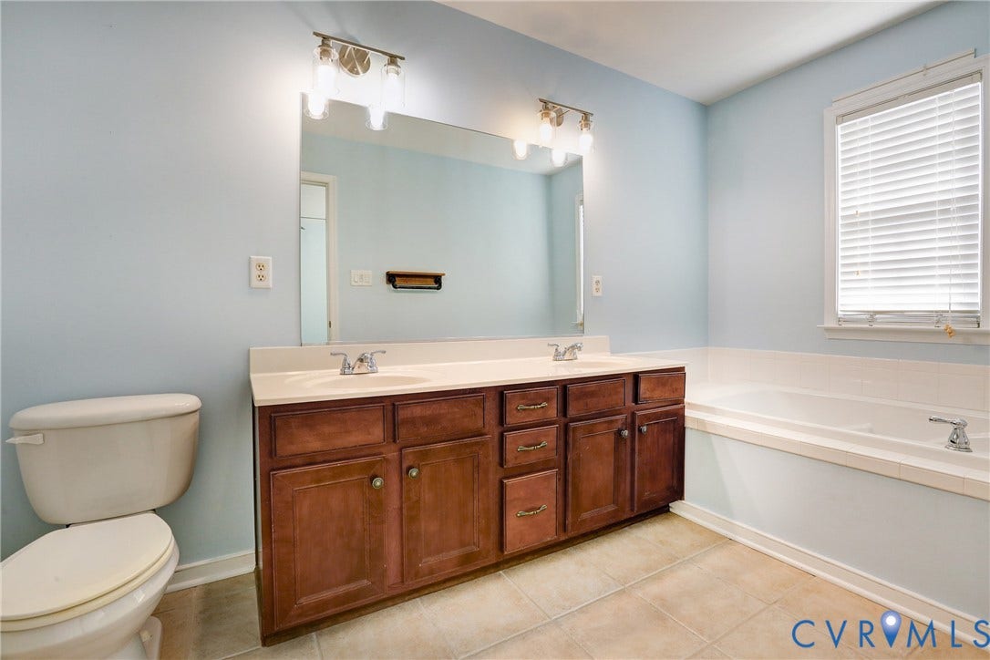 Bathroom featuring a wood-finish double vanity with an integrated countertop, large wall-mounted mirror, dual vanity lights, and a deep soaking tub with tile surround