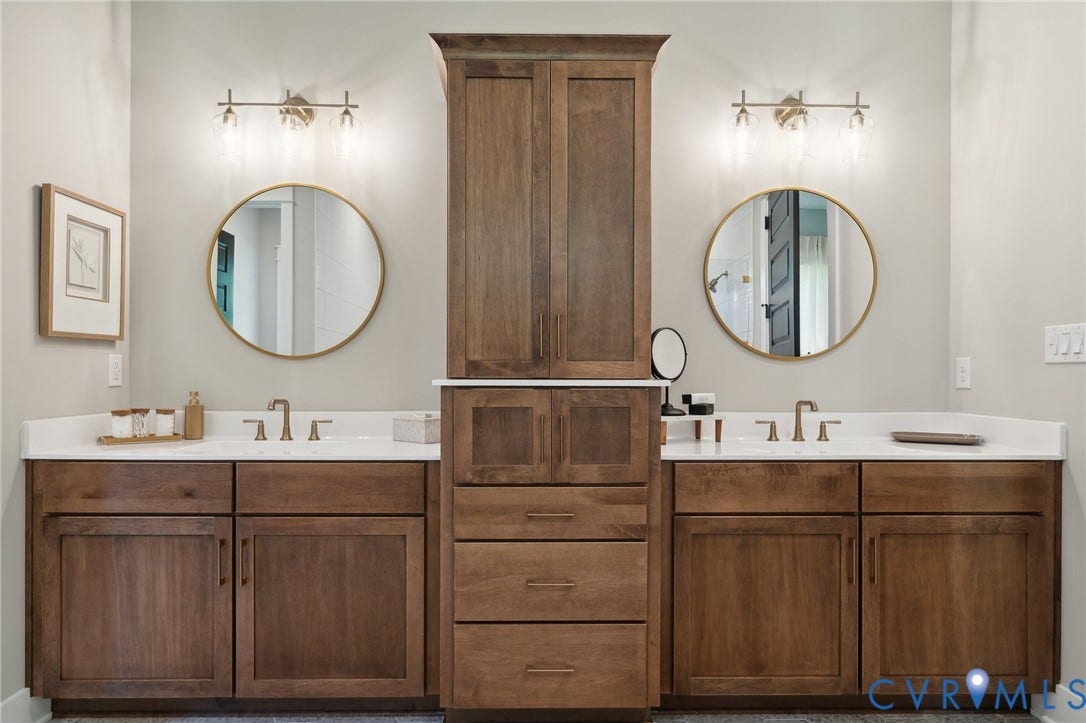 18128 Dogwood Trail Way, Rockville, VA 23146 Double vanity featuring wood-finish cabinetry, central tower storage, white countertops, dual circular mirrors with gold-tone frames, and three-light wall-mounted fixtures