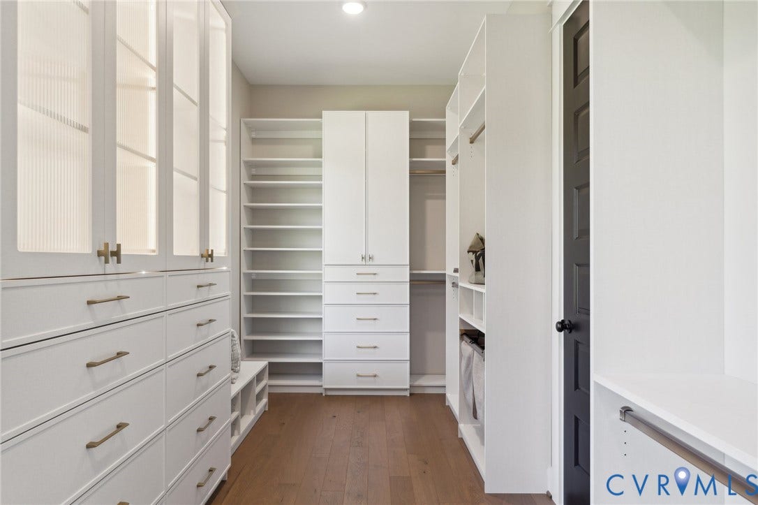 18128 Dogwood Trail Way, Rockville, VA 23146 Custom walk-in closet featuring white built-in cabinetry with frosted glass-front upper doors, soft-close drawers, brass hardware, wood-finish flooring, and a contrast black interior door