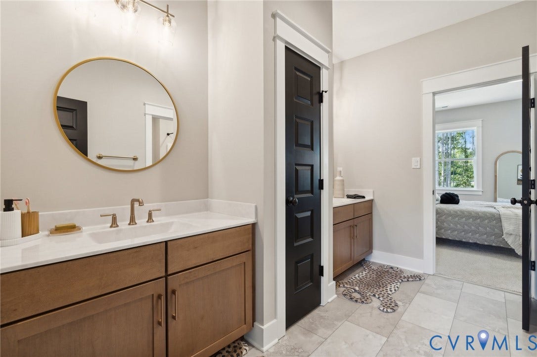 18128 Dogwood Trail Way, Rockville, VA 23146 Dual-vanity bathroom featuring light-finish cabinetry, solid-surface countertops, gold-toned circular mirror, and contrast black paneled doors