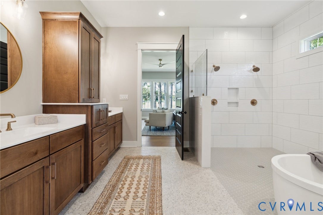 18128 Dogwood Trail Way, Rockville, VA 23146 Bathroom featuring wood-finish cabinetry, white solid-surface countertops, a walk-in shower with white tile surround and dual brass-finish showerheads, and a freestanding soaking tub