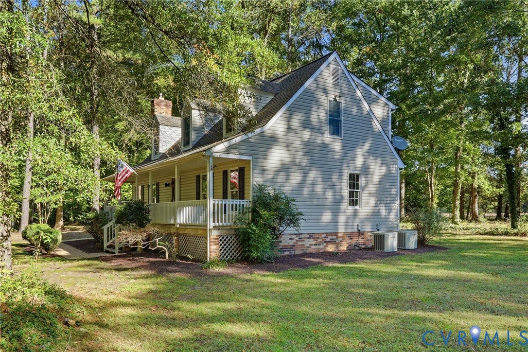 7270 Crossing Oaks Trail, Mechanicsville, VA 23111 Two-story residence featuring light-colored horizontal siding, a covered wrap-around porch with railing, dormer windows, a brick foundation base, and a sprawling lot with mature trees