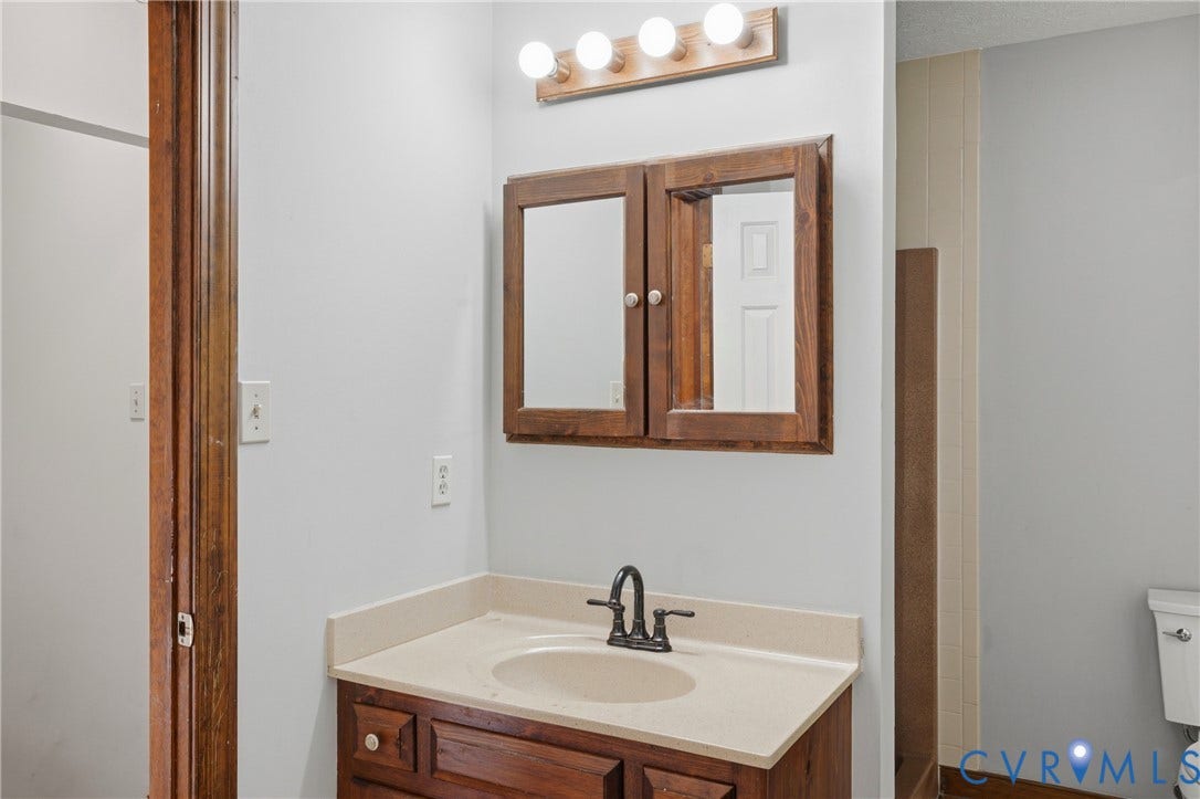 7270 Crossing Oaks Trail, Mechanicsville, VA 23111 Wood-finish vanity cabinet, solid-surface countertop with integrated basin, dark metal widespread faucet, wood-framed medicine cabinet, and four-light vanity fixture