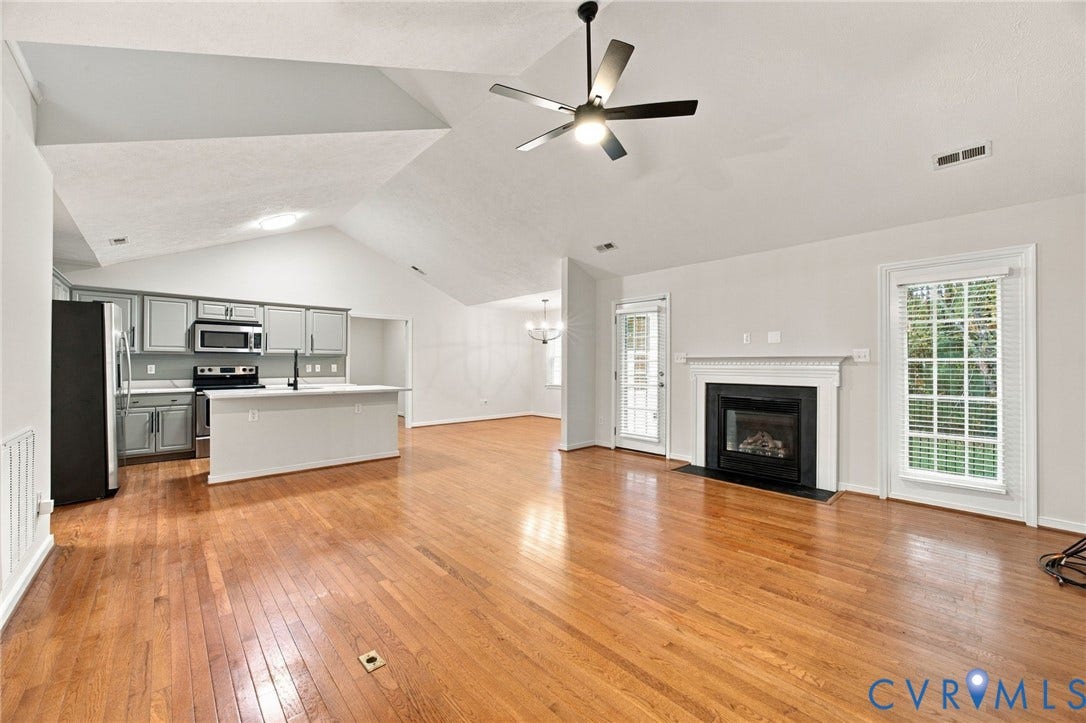 Vaulted ceilings featuring a central overhead fan, wood-finish flooring, a fireplace with a white mantel, kitchen cabinetry with a built-in microwave, and a peninsula counter