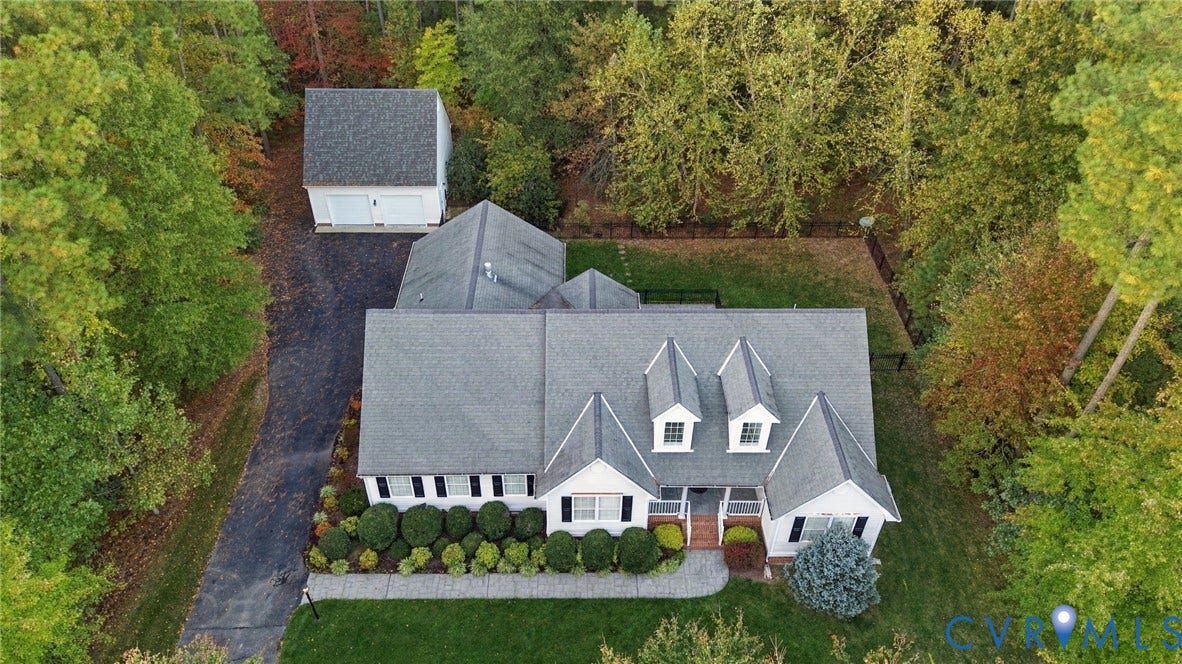 Aerial perspective of a detached residence featuring a dual-gable roofline, dormer windows, a separate two-car garage, and a paved driveway, all situated on a wooded lot