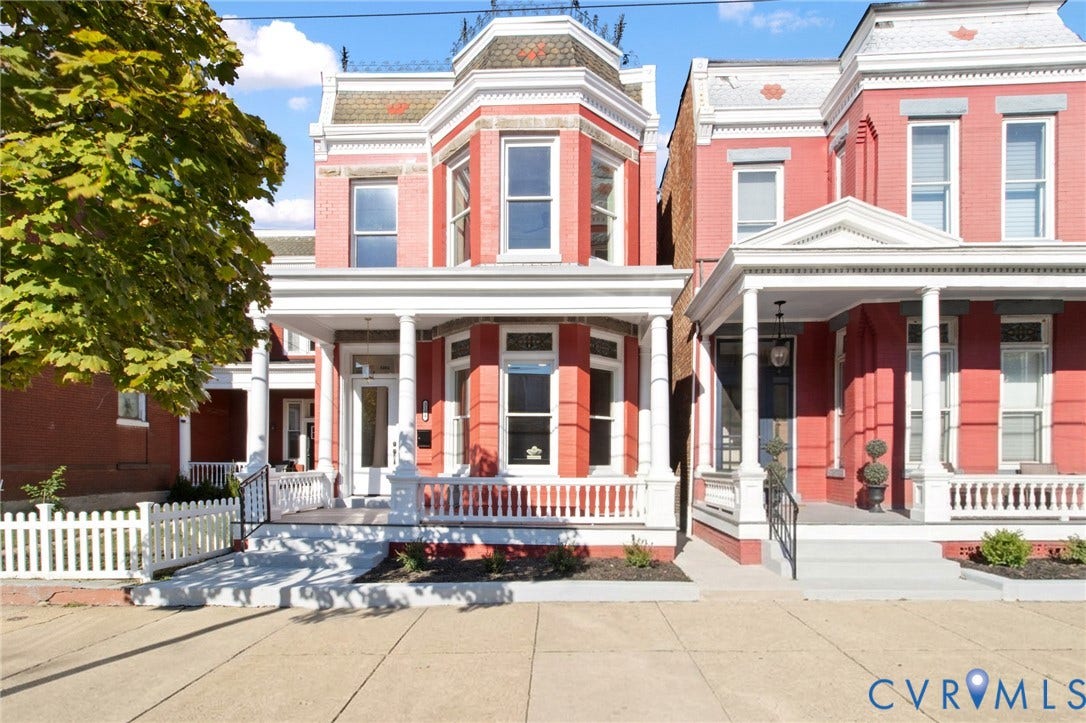 3104 E Broad Street, Richmond, VA 23223 Brick exterior featuring a two-story bay window, covered front porch with decorative columns, mansard-style roofline with shingles, and a white picket fence