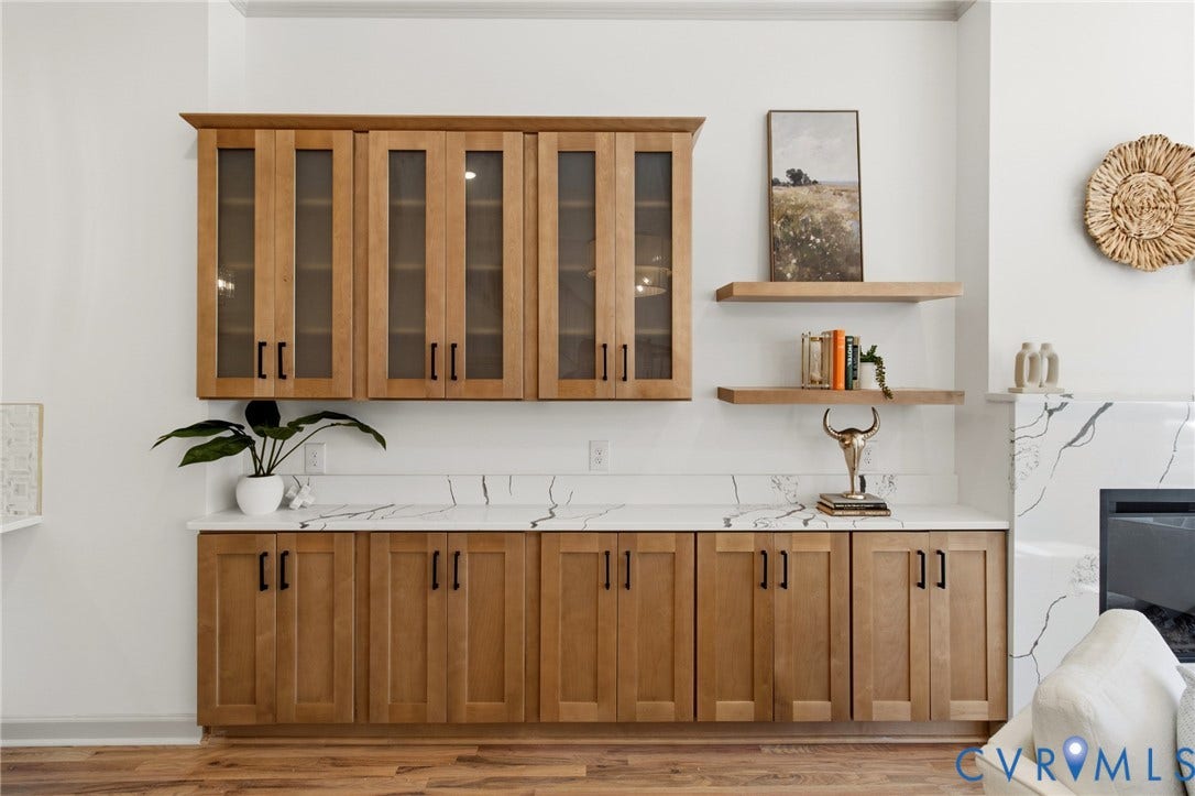 3104 E Broad Street, Richmond, VA 23223 Built-in cabinetry with wood-finish shaker doors, glass-front upper cabinets, stone-surfaced countertops with continuous matching backsplash, and custom wall-mounted floating wood shelves