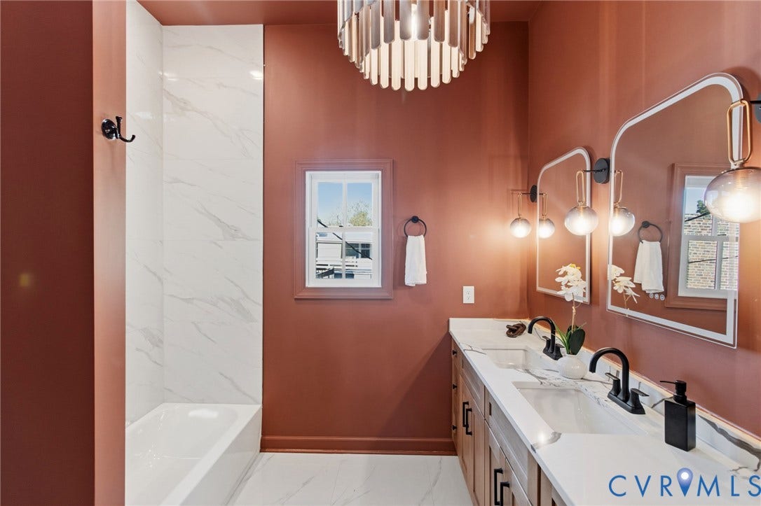 3104 E Broad Street, Richmond, VA 23223 Dual-sink vanity with light-tone wood cabinetry, white stone-finish countertops, matte black plumbing fixtures, wall-mounted mirrors with metallic frames, and floor-to-ceiling stone-finish shower surround