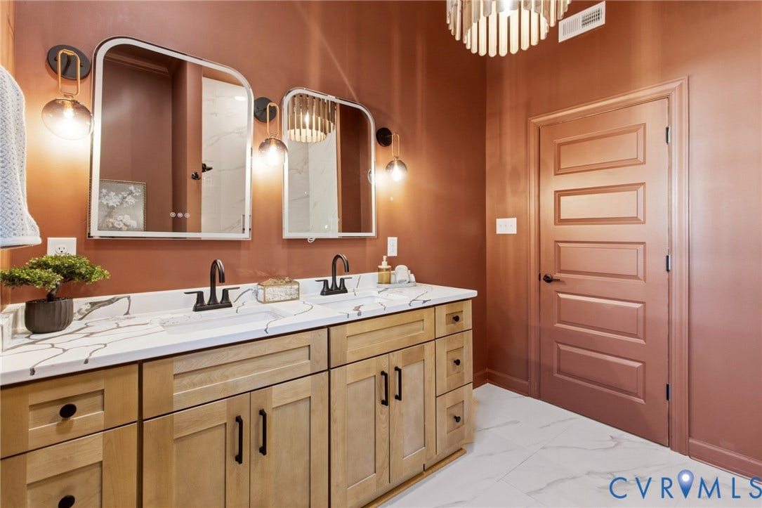 3104 E Broad Street, Richmond, VA 23223 Dual-sink vanity featuring light wood cabinetry, stone-surface countertop, and matte black hardware, paired with dual arched wall mirrors, decorative wall sconces, and white tile flooring