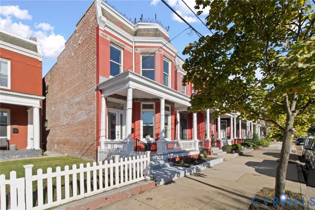 3104 E Broad Street, Richmond, VA 23223 Brick townhouse exterior featuring a covered front porch with classical columns, decorative roofline cresting, and a white picket fence