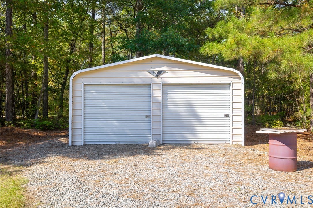 11400 County Drive, Prince George, VA 23842 Detached two-car metal garage featuring dual roll-up doors and a gravel driveway set against a wooded backdrop