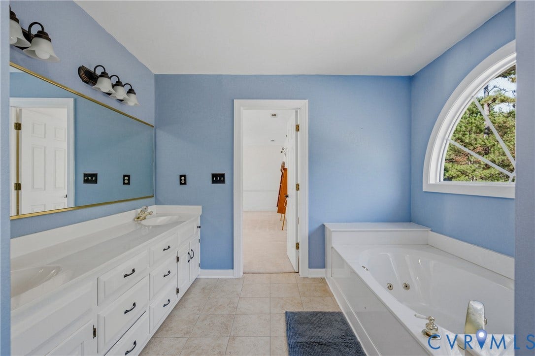 11400 County Drive, Prince George, VA 23842 Bathroom featuring a dual-sink vanity with white cabinetry, a large wall-mounted mirror with integrated lighting, a built-in soaking tub, and an arched window