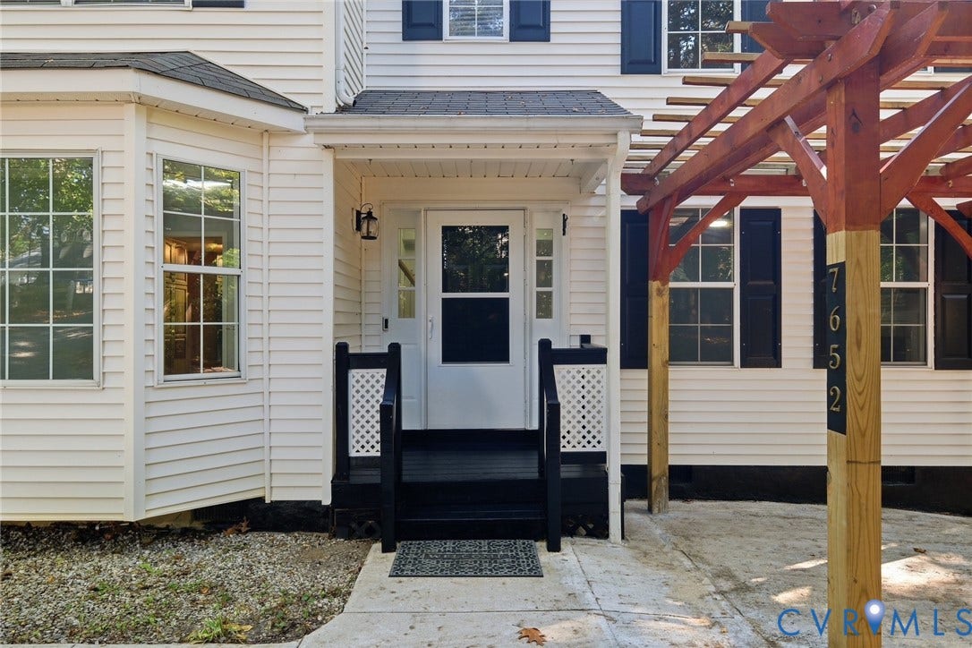 7652 Thacher Drive, Toano, VA 23168 Exterior entrance featuring white siding, black shutters, a covered porch with lattice-accented railings, and a wood-frame pergola
