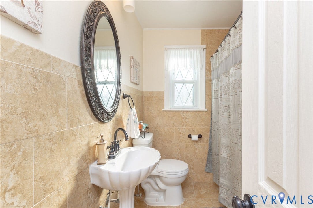 5211 W Grace Street, Richmond, VA 23226 Pedestal sink with oil-rubbed bronze faucet, neutral stone-finish wall and floor tiling, oval decorative mirror, and single-hung window with diamond muntins
