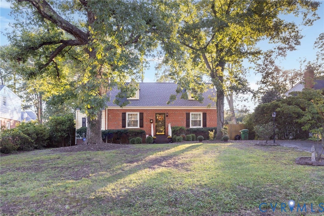 5211 W Grace Street, Richmond, VA 23226 Brick exterior residence featuring a gabled roofline, two dormer windows, and black shutters, situated on a lot with mature trees