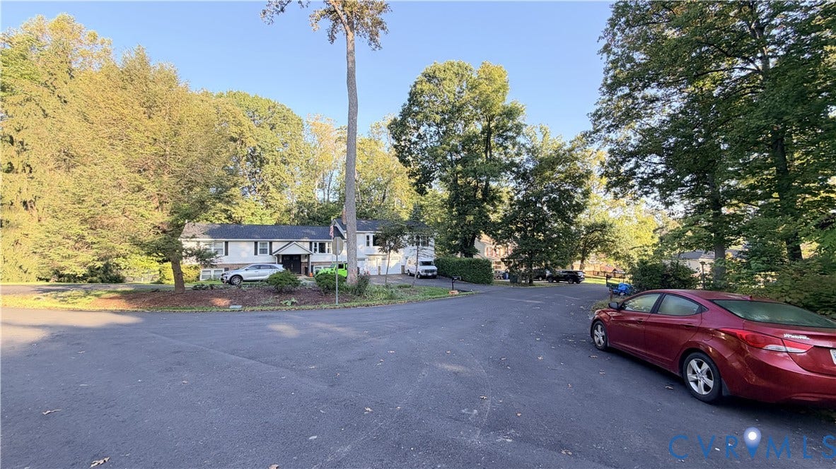 Two-story residence featuring light-colored siding, a multi-car garage, and mature trees on a lot with an expansive asphalt driveway