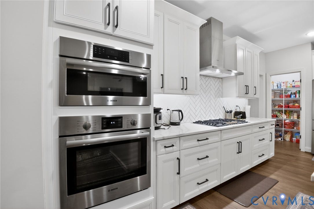 White cabinetry with matte black hardware, stainless steel built-in wall oven and microwave, stone-finish countertops, herringbone-patterned tile backsplash, and stainless steel range hood