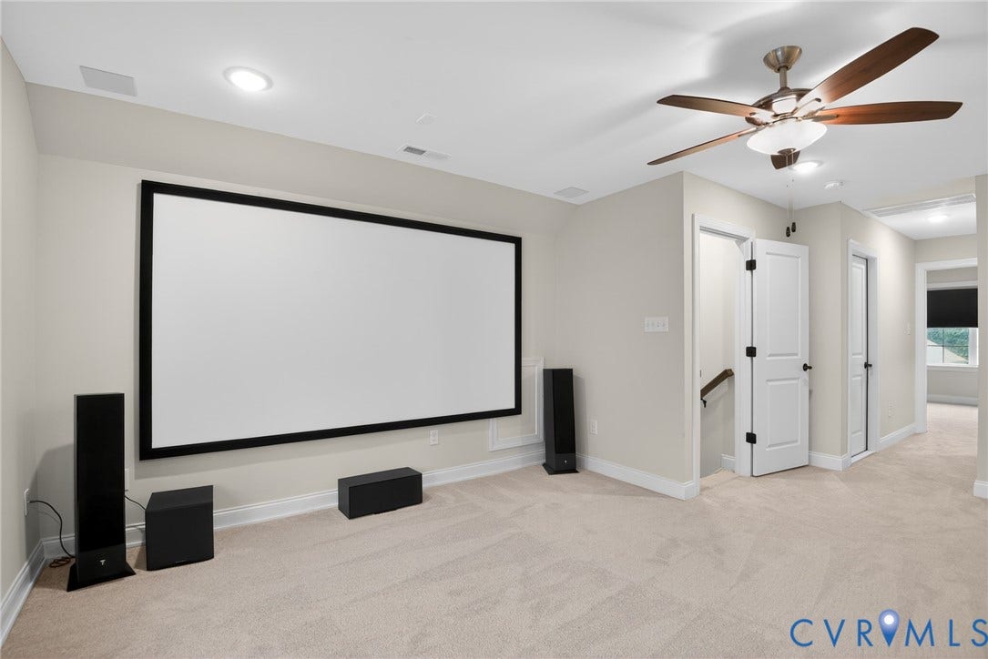 Dedicated space featuring neutral wall tones, wall-to-wall carpeting, a mounted projector screen, and a ceiling fan with integrated lighting