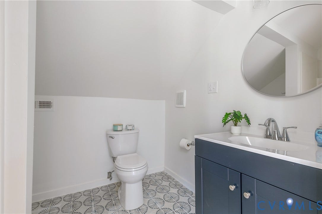 7263 Grace Lane, Mechanicsville, VA 23111 Bathroom featuring a white toilet, patterned floor tiles, a dark blue vanity with silver hardware, a white countertop with an integrated sink, and a circular mirror