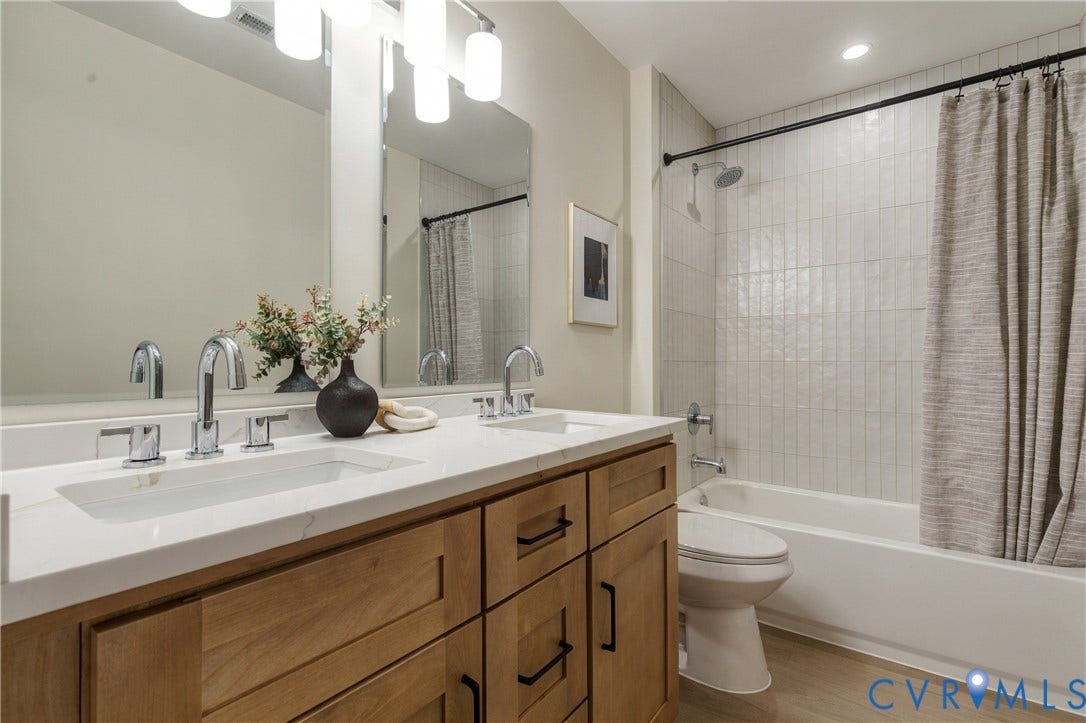 8060 Pebblepath Parkway, Hanover, VA 23111 Dual-sink vanity with light-toned wood cabinetry, stone-finish countertops, wall-mounted light fixtures, and a tub-shower combination featuring vertically-laid tile surround