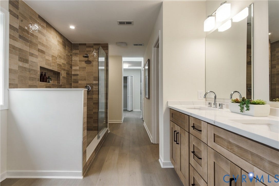 8060 Pebblepath Parkway, Hanover, VA 23111 Double vanity featuring wood-finish cabinetry, white countertop with dual undermount sinks, wall-mounted light fixtures, and glass-enclosed walk-in shower with textured tile wall and built-in recessed niche