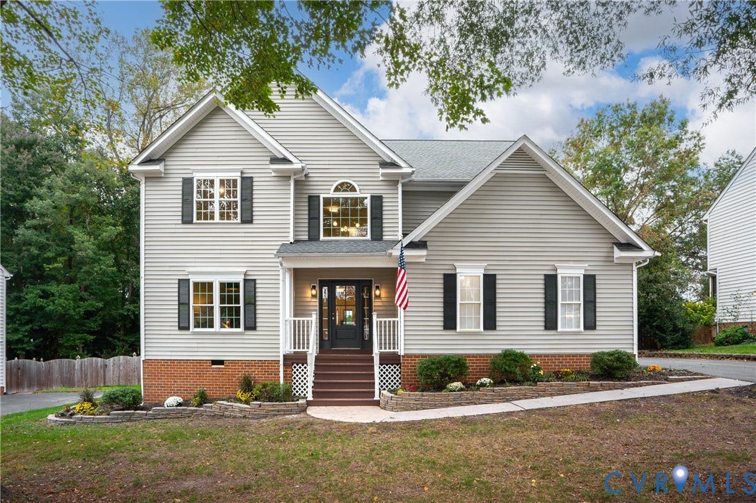 8060 Pebblepath Parkway, Hanover, VA 23111 Two-story residence featuring light gray horizontal siding, brick foundation facade, multi-pane windows with black shutters, covered front porch with a wood-finished staircase, and tiered stone garden wall
