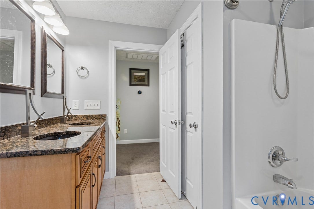Dual-sink vanity with stone-finish countertop, undermounted basins, and light wood-finish cabinetry, paired with a shower-tub combination featuring a handheld fixture and tiled flooring