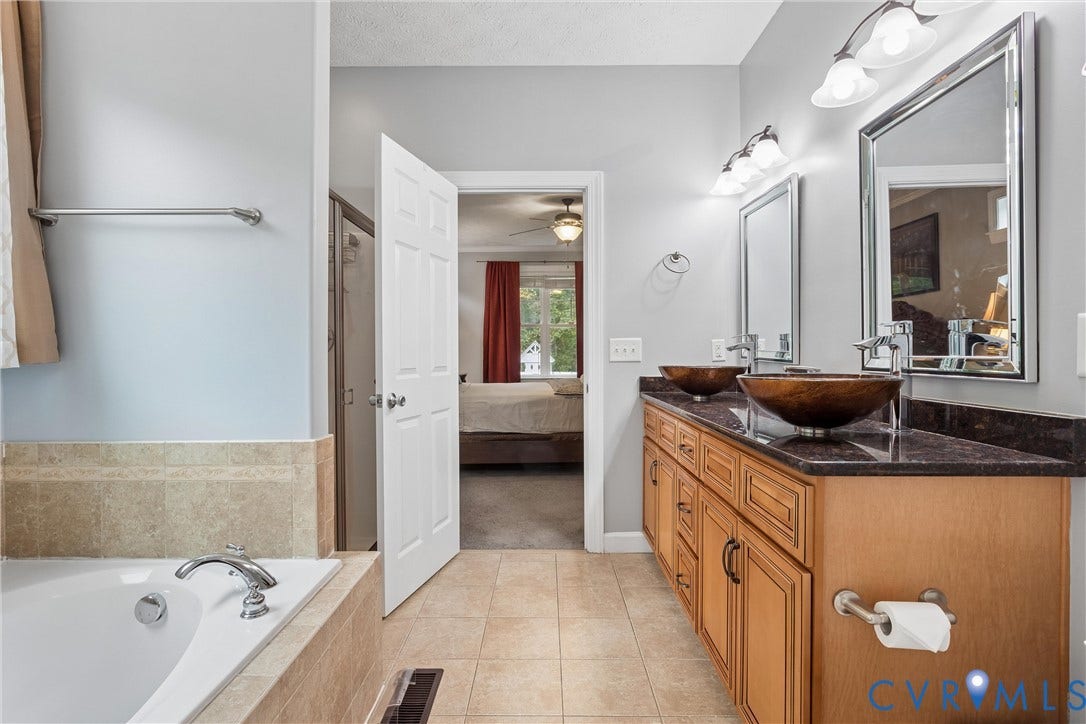 Dual-sink vanity featuring amber-toned vessel basins, dark stone-finish countertops, warm wood cabinetry, and framed mirrors with overhead light fixtures, paired with a built-in soaking tub finished in neutral tile