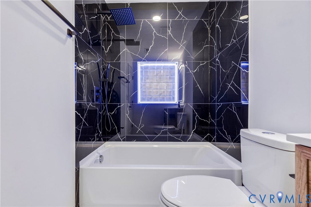 223 S Crater Road, Petersburg, VA 23803 Bathtub surround featuring black patterned tile, rainfall showerhead, handheld shower fixture, and a glass block window with integrated illumination